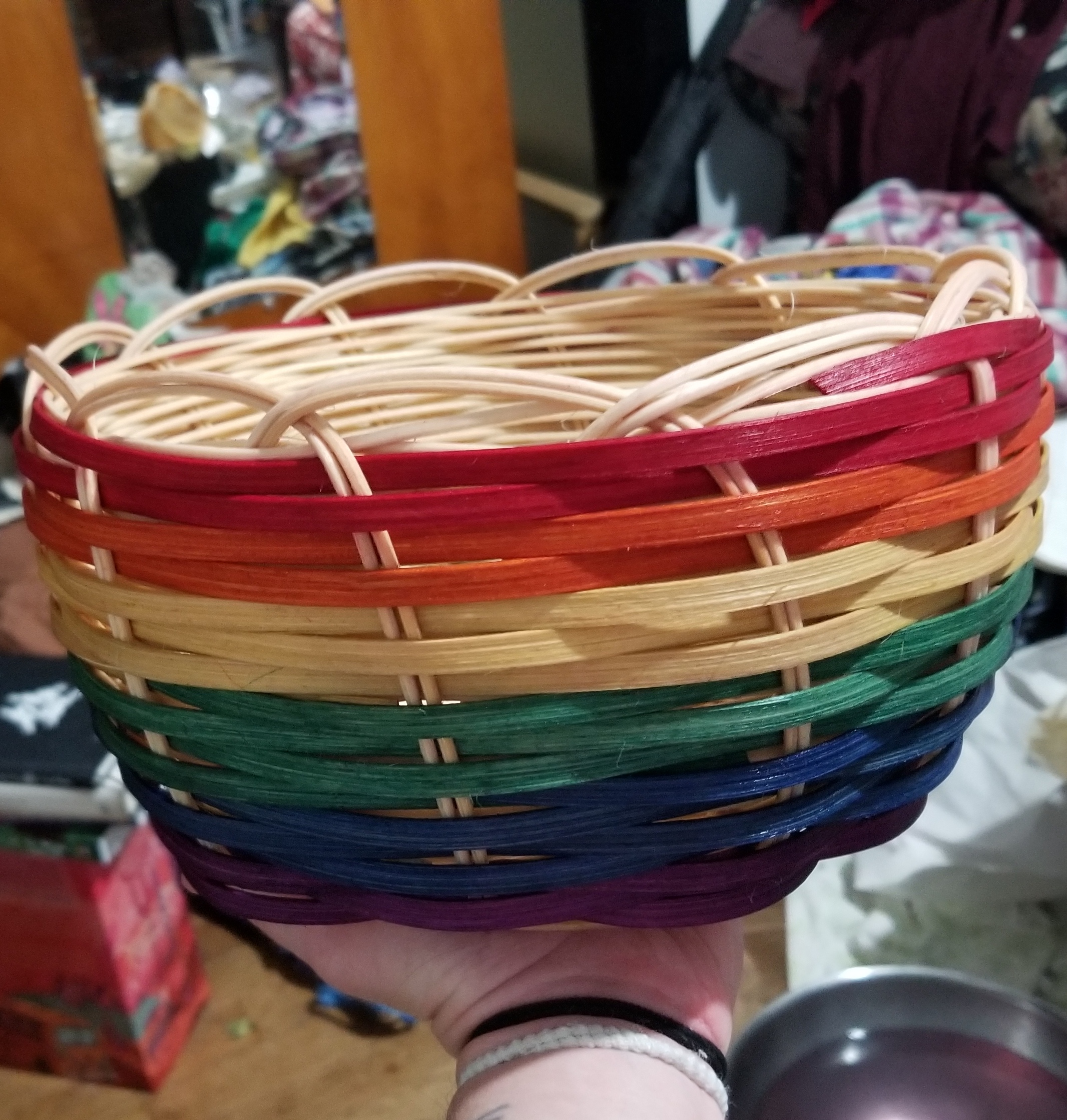 A large, circular, double-walled basket. The inner wall is natural-colored reed. The outside is colored flat cane in a rainbow pattern.